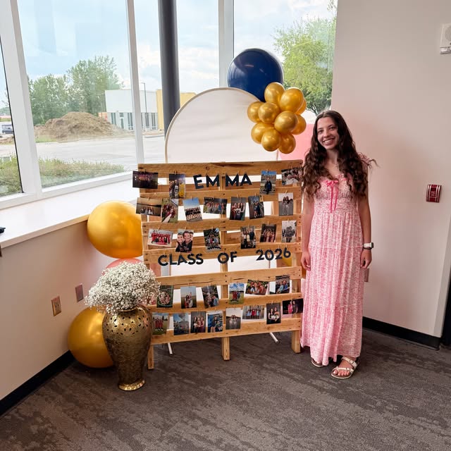 Emma standing beside a personalized graduation photo display made from a wooden pallet, with balloons and florals