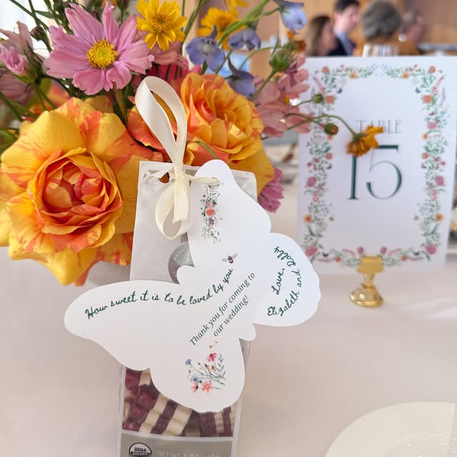 Butterfly-shaped personalized wedding favor tag beside colorful flowers and a Table 15 sign